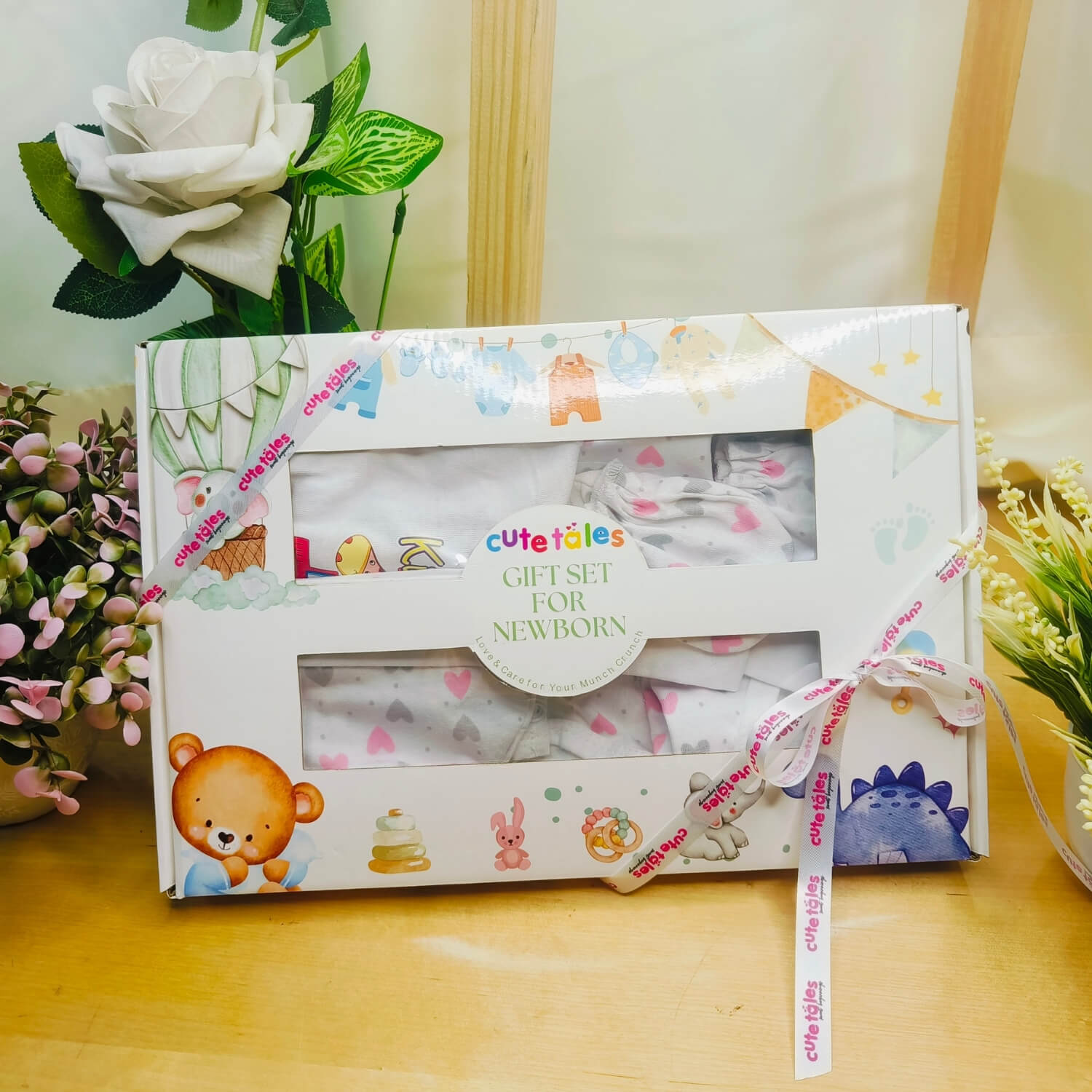 Cutetales gift set for newborn in a decorative box on a wooden surface with flowers in the background